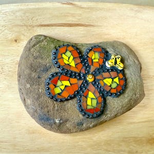 May include: A mosaic flower design on a natural stone. The flower features orange, yellow, and red glass tiles with a black border. A small frog figurine is attached to the stone. The stone is set on a wooden surface.