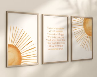 Op de afbeelding: Set van drie ingelijste kunstprints met aquarel zonontwerpen in oranje en gele tinten. Eén print toont de tekst "You are my sunshine". De frames hebben een lichte houten afwerking.