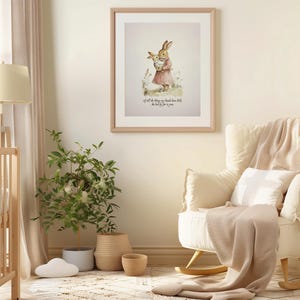 May include: A framed watercolor print of a mother rabbit holding a baby rabbit, in a nursery setting. The print is in a light wood frame. A white rocking chair with a beige blanket and pillow is in the foreground. A crib and a potted plant are also visible.