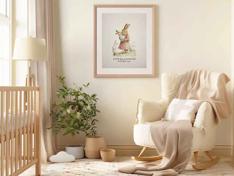 May include: A framed watercolor print of a mother rabbit holding a baby rabbit, in a nursery setting. The print is in a light wood frame. A white rocking chair with a beige blanket and pillow is in the foreground. A crib and a potted plant are also visible.