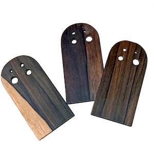Op de afbeelding: Drie platte, rechthoekige houten stukken met afgeronde bovenkanten, elk met een opvallend donkerbruin en beige houtnerfpatroon. Elk stuk bevat drie cirkelvormige gaten van verschillende groottes nabij de bovenrand, gerangschikt in een patroon.