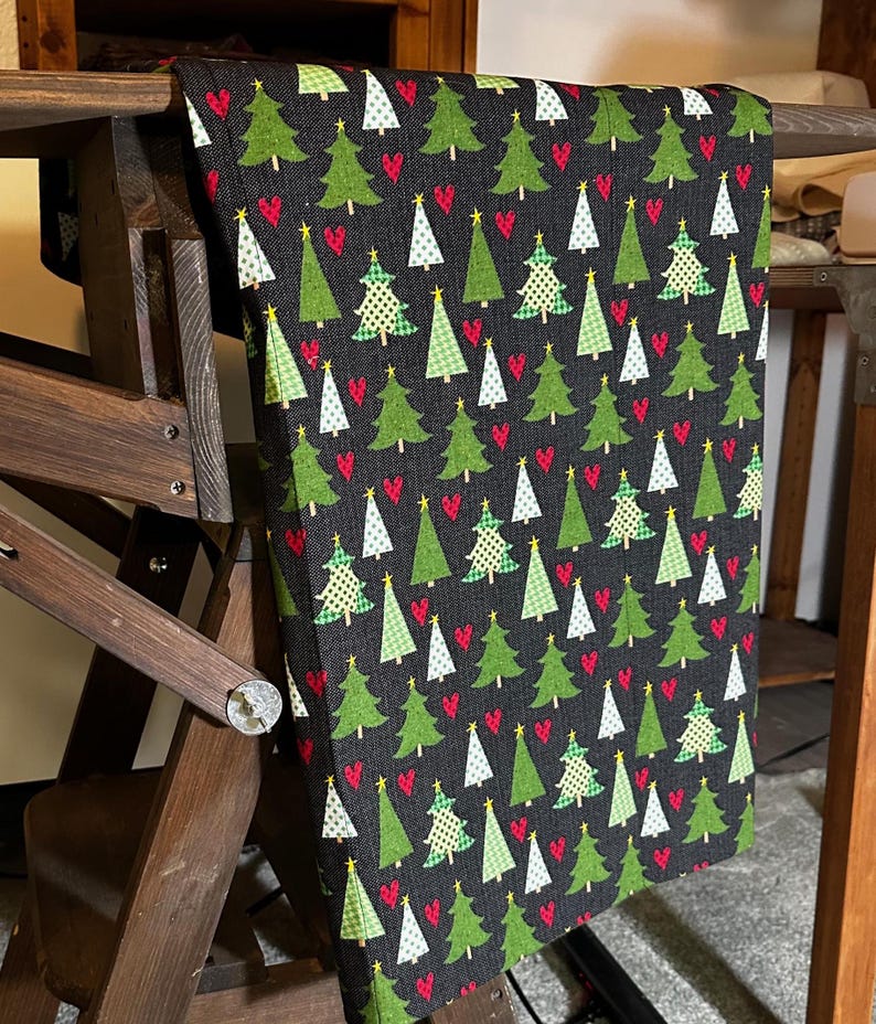May include: Black fabric with a repeating pattern of green Christmas trees and red hearts. The fabric is folded and draped over a wooden chair.