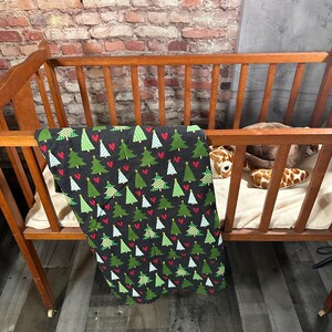 May include: A black and green crib blanket with a pattern of Christmas trees and red hearts. The blanket is hanging over the side of a wooden crib.