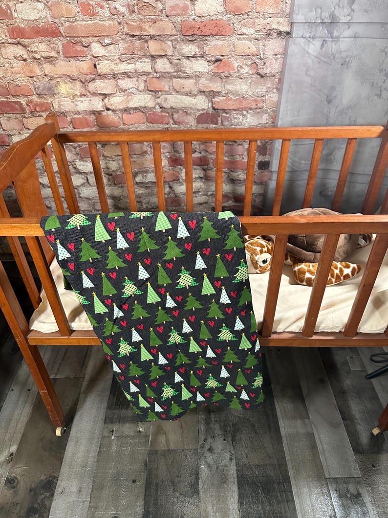 May include: A black and green crib blanket with a pattern of Christmas trees and red hearts. The blanket is hanging over the side of a wooden crib.