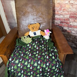 May include: A brown leather armchair with a green and red Christmas tree patterned blanket. A brown teddy bear wearing a blue shirt with the letter "B" on it is sitting on the blanket. There is a pink and white stuffed animal next to the bear.