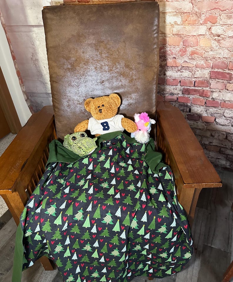 May include: A brown leather armchair with a green and red Christmas tree patterned blanket. A brown teddy bear wearing a blue shirt with the letter "B" on it is sitting on the blanket. There is a pink and white stuffed animal next to the bear.