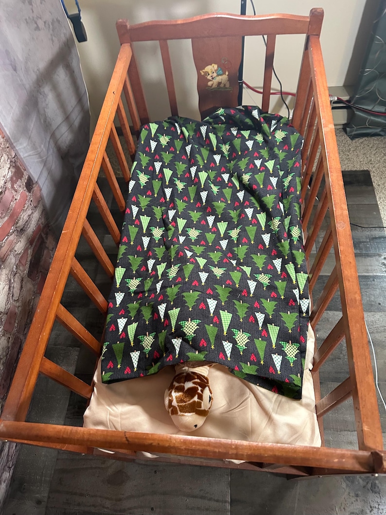May include: A wooden crib with a white and green Christmas tree patterned blanket and a stuffed giraffe toy inside.