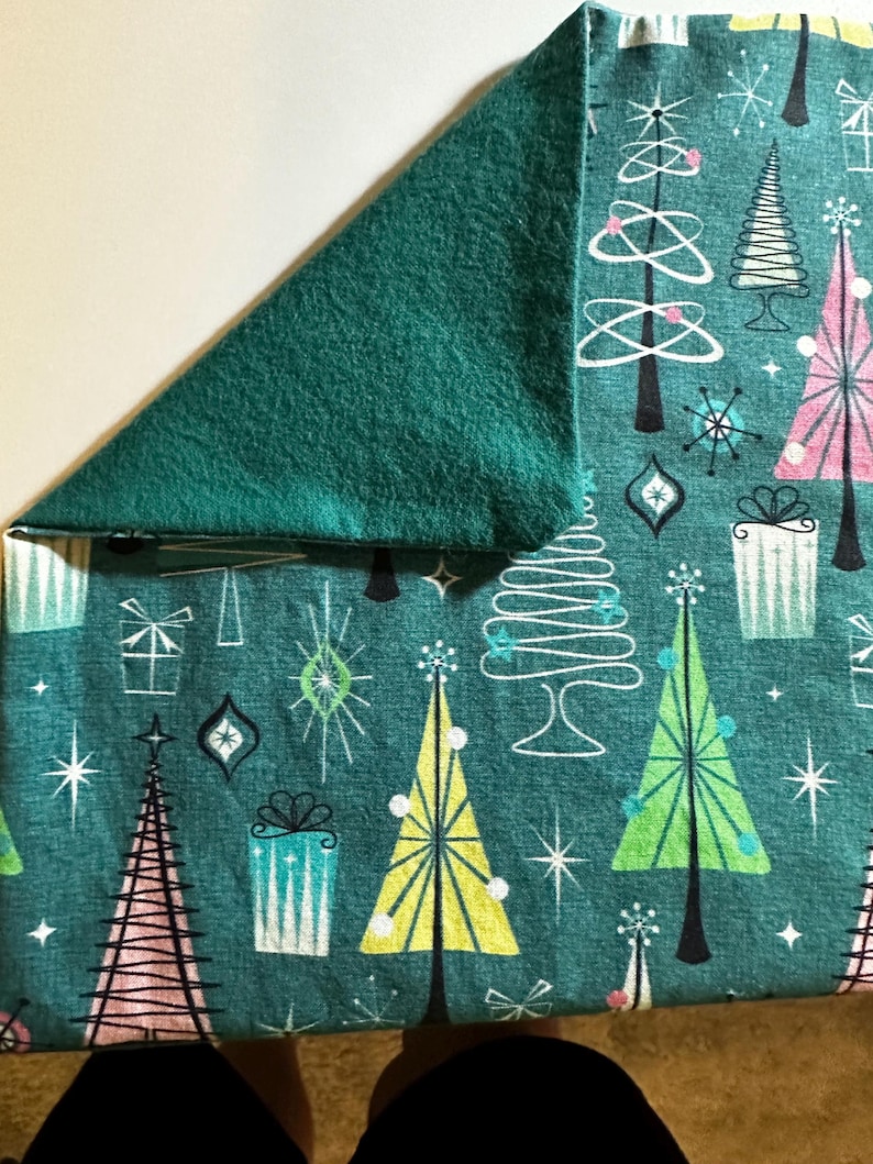 Puede incluir: Tela turquesa con un patr&oacute;n de &aacute;rboles de Navidad estilizados, estrellas y regalos. La tela tiene un respaldo de franela verde.