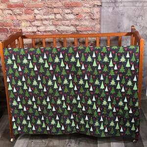 May include: A wooden crib with a black and green patterned crib sheet featuring a repeating pattern of Christmas trees and red hearts.