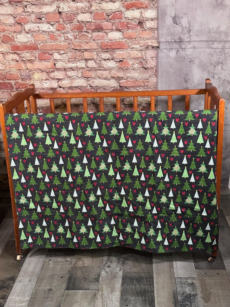 May include: A wooden crib with a black and green patterned crib sheet featuring a repeating pattern of Christmas trees and red hearts.