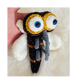 Op de afbeelding: Een handgemaakt, gehaakt insectenspeelgoed met grote, expressieve ogen. Het insect heeft een donkerbruin lichaam met gele en bruine strepen, witte vleugels en grijze poten. De ogen zijn wit met blauwe irissen en gele en bruine accenten.