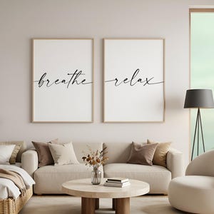 May include: Two framed prints with the words "breathe" and "relax" in cursive script. The prints hang above a beige sofa with cushions, creating a minimalist aesthetic. The decor is suitable for a living room or bedroom.