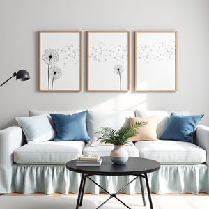 May include: Three vertical framed wall art prints featuring black line drawings of dandelion seed heads on a white background. The artwork is displayed in light wood frames, arranged in a row above a white sofa with blue and peach throw pillows.