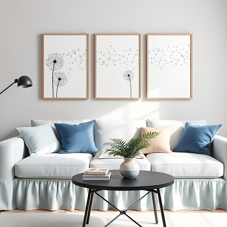May include: Three vertical framed wall art prints featuring black line drawings of dandelion seed heads on a white background. The artwork is displayed in light wood frames, arranged in a row above a white sofa with blue and peach throw pillows.