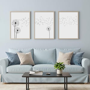 May include: A set of three rectangular wall art prints in light wood frames, each featuring a black and white line drawing of dandelion seed heads with seeds blowing across the white background, displayed in a horizontal row on a wall.