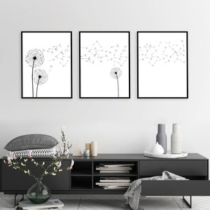 May include: A set of three framed wall art prints featuring black and white line drawings of dandelion seed heads. The artwork displays dandelion flowers with seeds blowing in the wind across a white background, enclosed in thin black frames.