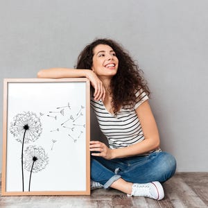 May include: A rectangular wooden picture frame containing a white print of two black dandelion heads with seeds blowing in the wind. The frame is positioned upright on a floor, showcasing a clean, minimalist botanical illustration in black ink.