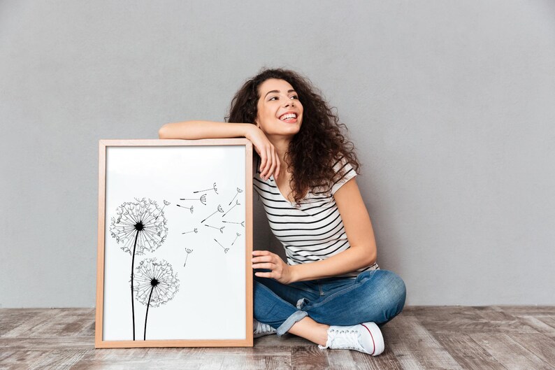 May include: A rectangular wooden picture frame containing a white print of two black dandelion heads with seeds blowing in the wind. The frame is positioned upright on a floor, showcasing a clean, minimalist botanical illustration in black ink.