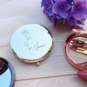 Peut inclure: Un miroir de poche doré avec un motif floral et le nom "Jean" en écriture manuscrite. Le miroir est rond et à charnière. Un autre miroir compact rose doré ouvert et un rouge à lèvres sont également visibles.