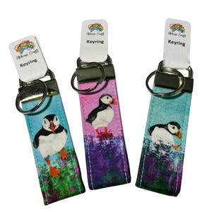 May include: Three fabric keyrings featuring a puffin illustration on a colourful background. Each keyring has a silver metal split ring and a white tag at the top with the text "Uphense Crafts Keyring" printed in black. The fabric designs show puffins.