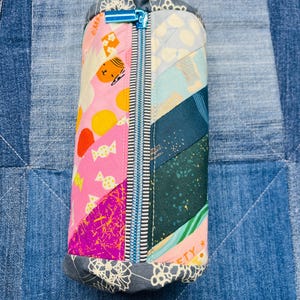 Peut inclure: Une trousse à crayons en patchwork coloré avec une fermeture éclair bleue. La trousse est faite de différents tissus avec des motifs variés, notamment rose, vert, bleu et blanc.
