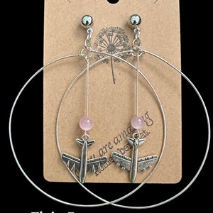 May include: Silver hoop earrings with a pink stone and a silver airplane charm. The earrings are displayed on a brown card with the text "You are amazing, remember that." and "Fly'n Betty Designs".