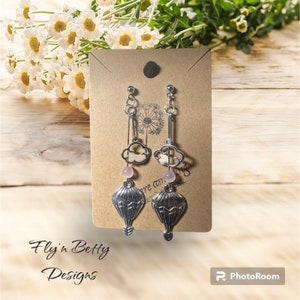 May include: A pair of silver earrings with a hot air balloon charm, a pink bead, and a cloud charm. The earrings are displayed on a brown card with the text "Fly 'n Betty Designs".