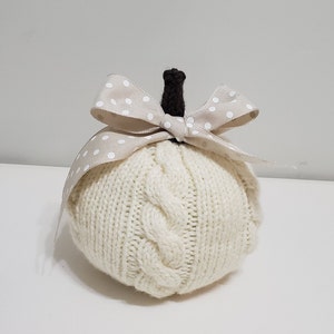 May include: A white knitted pumpkin with a brown stem and a beige bow with white polka dots.