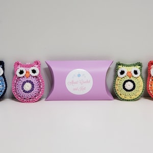May include: Five crocheted owl keychains in different colors, including blue, pink, green, and orange. The owls have black eyes and a white circle in the center of their bodies. The keychains are packaged in a purple pillow box with a white label that says "Meet Crocheted and Knit".