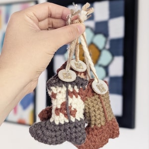 Puede incluir: Tres pequeños adornos de bota de ganchillo en tonos grises, marrones y crema, sujetos por hilo natural. Cada bota tiene una pequeña etiqueta redonda de madera con caracteres chinos dorados. Las botas cuelgan frente a una obra de arte enmarcada.