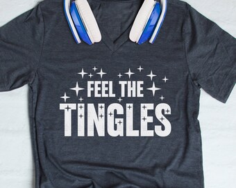 May include: A dark grey t-shirt with a white v-neck and the text "FEEL THE TINGLES" in white with white stars around the text. The t-shirt is lying flat on a white surface with a pair of white headphones on top of the shirt.