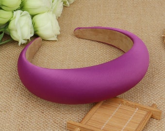 May include: A vibrant magenta satin headband with a padded design. The headband has a smooth, curved shape and a light brown interior. It is displayed on a textured, neutral-colored surface, with white flowers in the background.