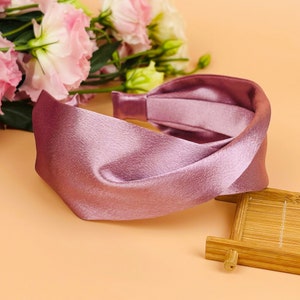 May include: A pink satin headband with a twisted knot design. The headband is lying on a light brown surface.