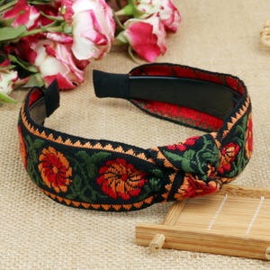 May include: A black headband with a floral pattern of red and orange flowers, green leaves, and an orange trim. The headband has a knotted design and a black interior.
