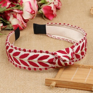 May include: Red and white headband with a knotted design and gold leaf embroidery. The headband is made of fabric and has a soft, comfortable feel.