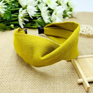 May include: Mustard yellow fabric headband with a twisted knot design. The headband has a black accent at the back and is made of a textured material. The accessory is displayed on a neutral background with white flowers.