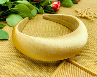 Könnte beinhalten: Ein breites, goldenes Satin-Haarband. Das Haarband ist dick und hat eine glatte, abgerundete Form.