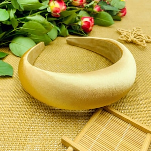 Könnte beinhalten: Ein breites, goldenes Satin-Haarband. Das Haarband ist dick und hat eine glatte, abgerundete Form.