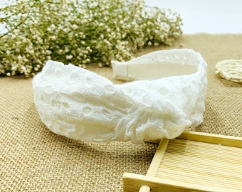 May include: White lace headband with a knotted design. The headband is on a burlap surface.