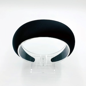 K&ouml;nnte beinhalten: Ein schwarzes Stirnband mit glatter, matter Oberfl&auml;che. Das Stirnband wird auf einem durchsichtigen Plastikst&auml;nder pr&auml;sentiert.