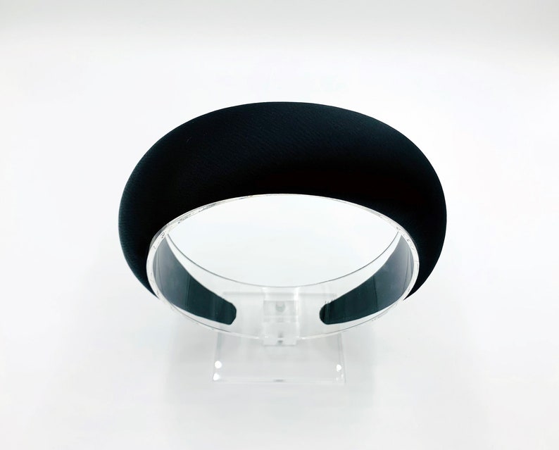 K&ouml;nnte beinhalten: Ein schwarzes Stirnband mit glatter, matter Oberfl&auml;che. Das Stirnband wird auf einem durchsichtigen Plastikst&auml;nder pr&auml;sentiert.