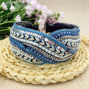 May include: A blue and white headband with a braided design and beaded embellishments. The headband is made of fabric and has a twisted knot design.