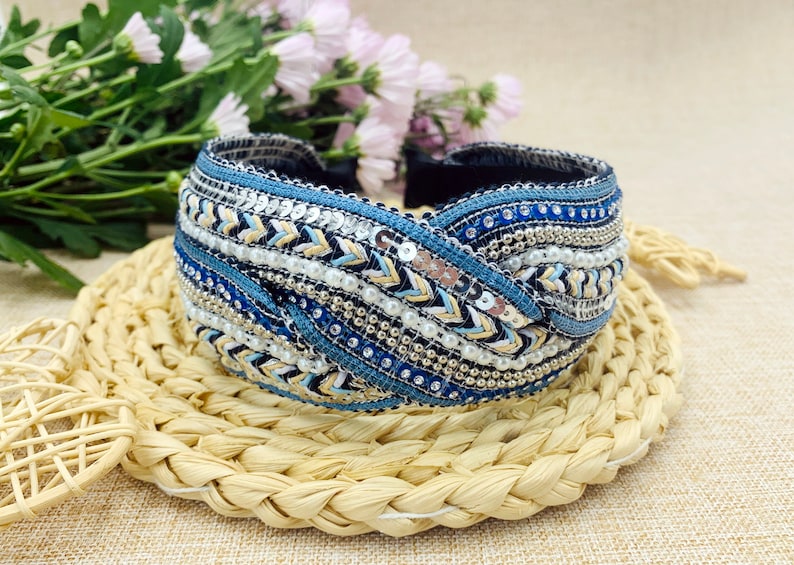 May include: A blue and white headband with a braided design and beaded embellishments. The headband is made of fabric and has a twisted knot design.