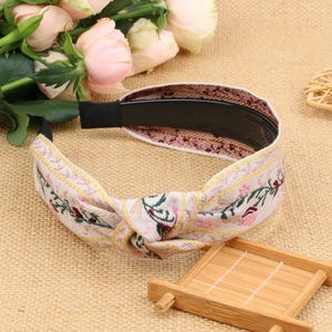 May include: A white headband with a floral pattern and a knotted design. The headband is made of fabric and has a black elastic band.