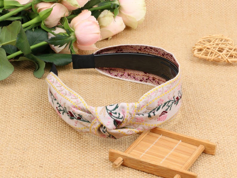 May include: A white headband with a floral pattern and a knotted design. The headband is made of fabric and has a black elastic band.