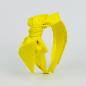 Op de afbeelding: Een felgeel hoofdband met een grote, oversized strik. De hoofdband is gemaakt van een zachte, satijnachtige stof.