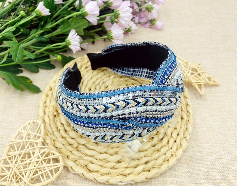 May include: A blue and white beaded headband with a knotted design. The headband is made of fabric and has a black elastic band.