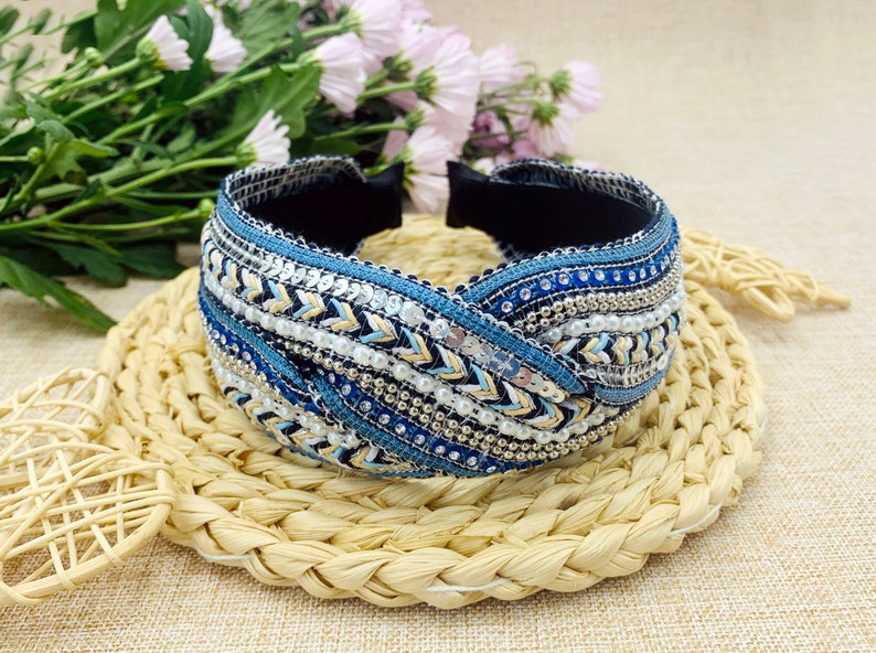 May include: A blue, white, and gold beaded headband with a braided design. The headband is made of fabric and has a variety of beads and sequins.