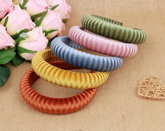 May include: A collection of colorful, ribbed velvet bracelets in shades of orange, yellow, blue, pink, and green. The bracelets are stacked on a neutral-toned surface, with a bouquet of pink roses in the background.