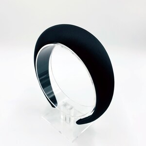 K&ouml;nnte beinhalten: Ein schwarzes Samt-Haarband mit einer breiten, abgerundeten Form. Das Haarband wird auf einem klaren Acryl-St&auml;nder pr&auml;sentiert.